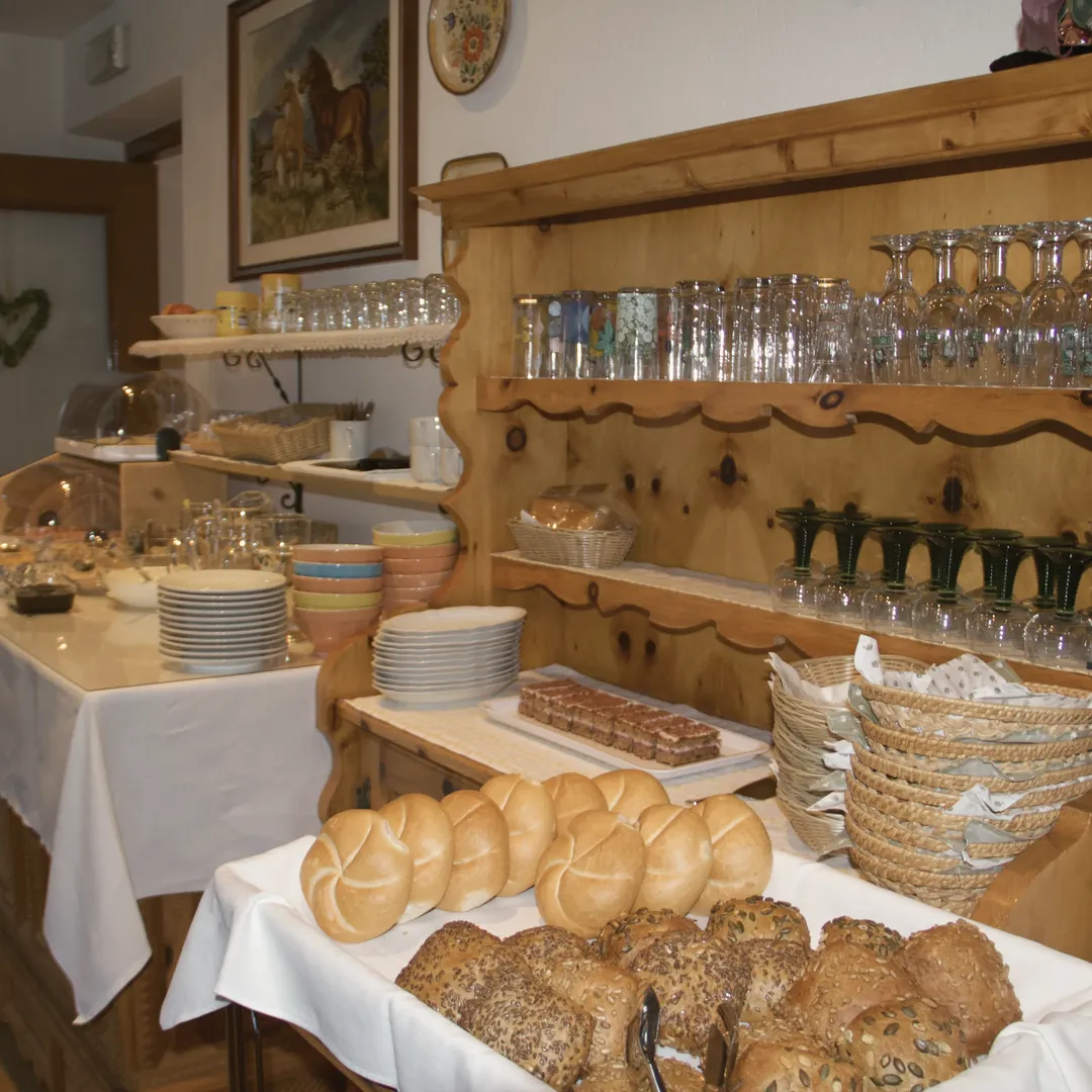 Il buffet della colazione con panini, pane ai cereali e dolce, davanti a una credenza in legno con i bicchieri